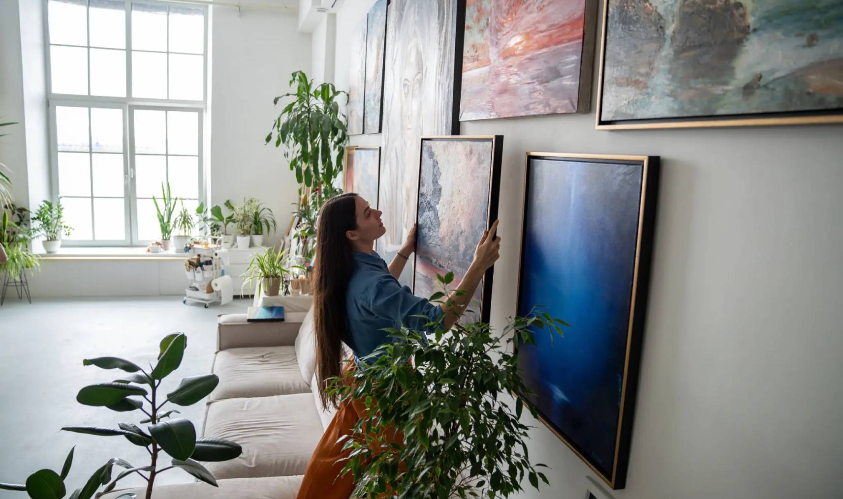 Woman hanging artwork in a stylish living room filled with plants.