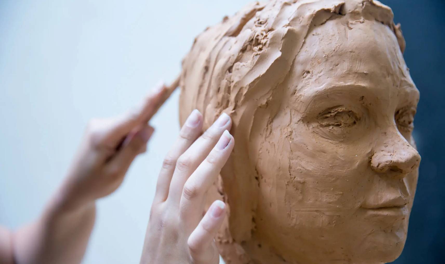 Hands sculpting a clay face with a wooden tool.